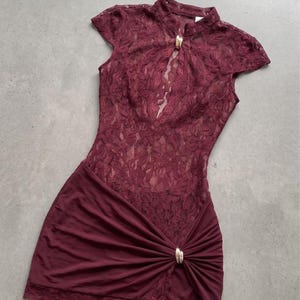 Puede incluir: Un vestido corto de color burdeos con un cuerpo de encaje floral, cuello alto y una abertura tipo cerradura asegurada con un broche de metal dorado. La falda est&aacute; confeccionada en tela lisa con un detalle fruncido en el lateral.