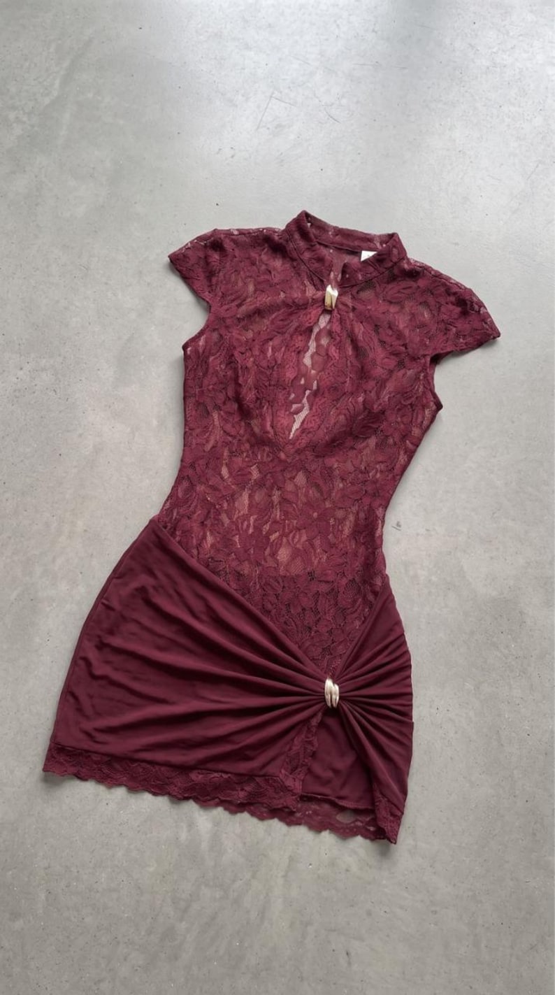 Puede incluir: Un vestido corto de color burdeos con un cuerpo de encaje floral, cuello alto y una abertura tipo cerradura asegurada con un broche de metal dorado. La falda est&aacute; confeccionada en tela lisa con un detalle fruncido en el lateral.