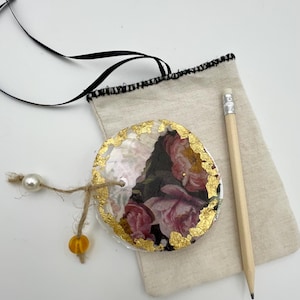 Op de afbeelding: Een ronde, witte, iriserende schelp met een bloemenpatroon en gouden accenten. De schelp hangt aan een touwtje met een witte parel en een gele kraal.