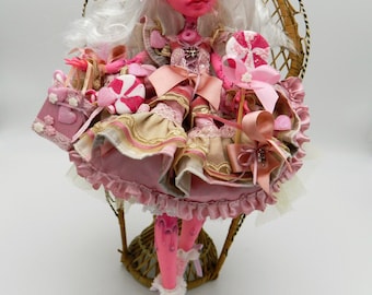 Peut inclure: Une poupée rose aux cheveux blancs portant une robe rose et dorée avec de la dentelle et des nœuds. La poupée est assise dans un fauteuil en osier et tient une sucette rose et blanche.