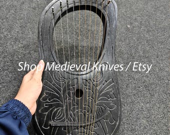 Może przedstawiać: Czarna, rzeźbiona lira ze strunami, z motywami kwiatowymi. Instrument ma zaokrąglony kształt z otworem dźwiękowym i jest trzymany przez osobę. Tekst "Shop Medieval Knives / Etsy" jest widoczny.