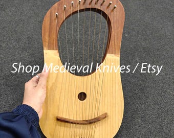 Może przedstawiać: Drewniana lira z jasnobrązowym korpusem i ciemnobrązowym, zakrzywionym wierzchem. Instrument posiada struny i otwór rezonansowy. Na instrumencie wydrukowano tekst "Shop Medieval Knives / Etsy".