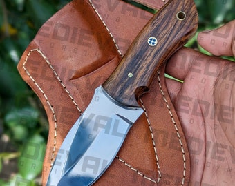 May include: A brown leather sheath with a stitched edge holds a small, fixed blade knife with a dark wood handle. The blade is made of polished steel and has a slight curve. The handle has a brass pin and a small cross design.
