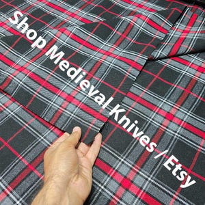 May include: Close-up of a black, red, and gray plaid fabric. The fabric has a repeating pattern of intersecting lines in red, gray, and white on a black background. The text "Shop Medieval Knives / Etsy" is printed in white.
