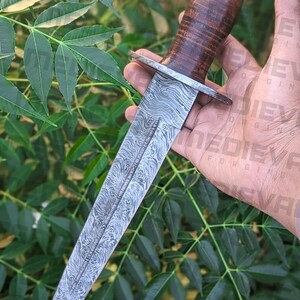 以下が含まれることがあります： 木製のハンドルが付いた銀の短剣。刃は模様があり、非常に鋭利です。短剣は緑の葉の背景に手を添えて持たれています。