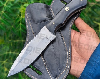 May include: A black handled knife with a silver blade and a leather sheath. The knife is held in a hand with the sheath in the background.