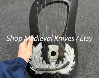 Może przedstawiać: Czarna lira ze srebrnymi elementami dekoracyjnymi i strunami. Instrument ma zaokrąglony kształt. Widoczny jest tekst "Shop Medieval Knives / Etsy". Lira jest trzymana przez osobę.