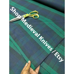 May include: A close-up of green and navy blue plaid fabric with gold scissors. The fabric is folded, and a hand is holding the corner. The text "Shop Medieval Knives / Etsy" is overlaid on the fabric.