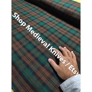 May include: A close-up of a plaid fabric in shades of brown, green, and black. The fabric has a traditional tartan pattern. The text "Shop Medieval Knives / Etsy" is printed across the fabric.