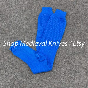 Op de afbeelding: Felblauw gebreide beenbeschermers. Het bovenste deel heeft een textuurpatroon, terwijl het onderste deel geribbeld is. De tekst "Shop Medieval Knives / Etsy" is zichtbaar.