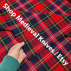 Puede incluir: Tela a cuadros roja, azul marino y amarilla con el texto "Shop Medieval Knives / Etsy" en blanco. La tela está siendo sostenida por una persona que lleva una sudadera verde.