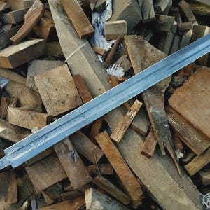 May include: A long, silver sword with a textured blade lies atop a pile of wooden planks. The sword's handle has a detailed pattern. The wood pieces vary in size and color, creating a rustic backdrop.