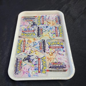 May include: A white rectangular tray holds six packs of Pokémon Trading Card Game: Scarlet & Violet - Prismatic Evolutions. The packs feature colorful graphics of Pokémon characters and the game's logo.