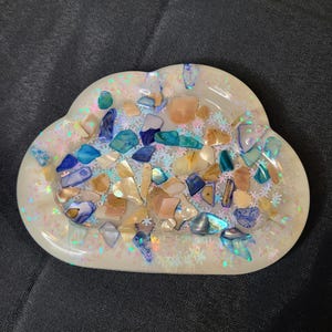 May include: Cloud-shaped ashtray with a pearlescent finish. The ashtray is filled with colorful, iridescent pieces of shell and stone in shades of blue, beige, and green. Snowflake-shaped glitter adds sparkle.