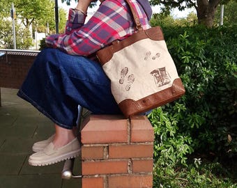 Puede incluir: Un bolso tote de lona crema con ribetes y asas de cuero marrón. La bolsa presenta huellas de pies y botas marrones sobre la lona. La bolsa es llevada por una persona que lleva una camisa a cuadros y vaqueros azules.