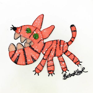 May include: A whimsical, orange sequined creature with black stripes and green eyes. The cartoon-like design features a large mouth with teeth and a curled tail. The artist's signature, "Barb Klor", is visible in the lower right corner.