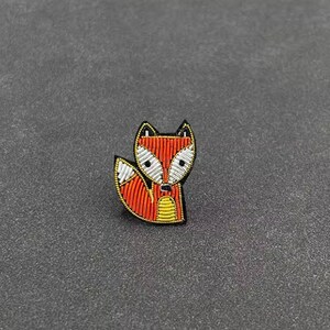 May include: A fox-shaped brooch crafted with intricate beadwork. The design features vibrant orange, white, and yellow beads arranged in rows to form the fox's face, ears, and bushy tail, all outlined with a thin black border for definition.