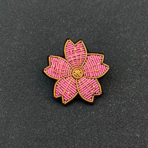 May include: A five-petaled flower brooch crafted with pink glass beads and metallic gold thread. The center features a circular gold-toned element, while the edges of each petal are outlined in gold wire, creating a detailed floral design.