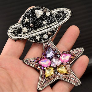 May include: A black and silver beaded planet with a star design, attached to a pink, purple, and yellow beaded star.