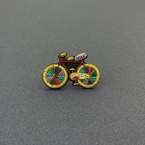 Peut inclure: Un écusson brodé en forme de vélo avec un cadre rouge, des roues à bordure dorée aux rayons multicolores bleus, verts, rouges et violets, un guidon jaune et une selle violette, le tout souligné par un fil noir sur une base en tissu sombre.