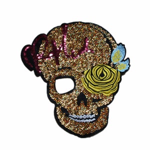 Puede incluir: Calavera de purpurina dorada con una rosa amarilla y una flor azul. La calavera tiene un diseño de purpurina rosa y dorada en la parte superior.