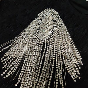 Puede incluir: Un adorno de flecos de pedrería plateada con un gran grupo de pedrería de forma ovalada en la parte superior.