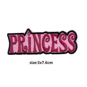 Pode incluir: Etiqueta de ferro bordada rosa e vermelha com a palavra "PRINCESA" em uma fonte estilizada. A etiqueta mede 3 cm por 7,6 cm.