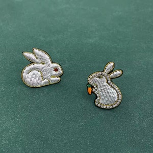 Peut inclure: Deux broches brodées en forme de lapin avec du fil métallique argenté et des bords dorés. Un lapin est assis de profil avec un œil rouge, tandis que l'autre est debout, tenant une petite carotte orange aux feuilles vertes et orné de strass.
