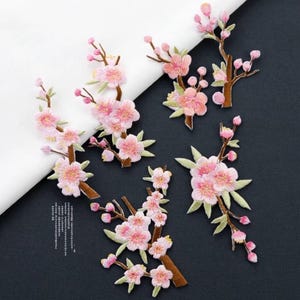May include: Four embroidered fabric patches featuring cherry blossom branches with light pink flowers, brown stems, and small green leaves. The patches are arranged on a surface that is partially white and partially dark grey.
