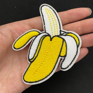 Puede incluir: Un parche de plátano amarillo bordado con contornos blancos y negros. El plátano está pelado y la cáscara está enrollada alrededor del plátano.