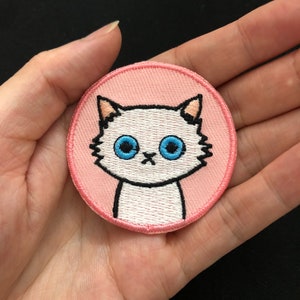 Puede incluir: Un parche redondo de color rosa con un gato blanco bordado con ojos azules.
