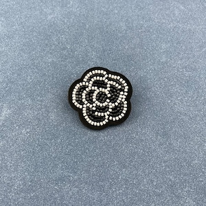 Può includere: Una spilla a forma di fiore con perline bianche e nere. La spilla presenta uno sfondo scuro con un motivo floreale realizzato con piccole perline bianche. Il fiore ha una forma arrotondata ed è centrato sulla spilla.