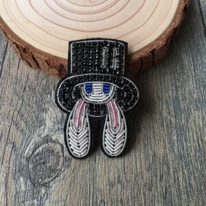 May include: A handcrafted brooch featuring a black top hat adorned with black crystals and silver metallic thread. The design includes a central face with blue and silver accents, flanked by two long, downward-pointing ears made of silver and pink thread.
