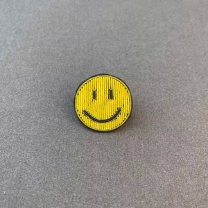 May include: A circular pin featuring a yellow smiley face design. The face is constructed from vertical yellow beads or thread-like textures, with two black vertical rectangles for eyes and a curved black line forming a wide, smiling mouth.
