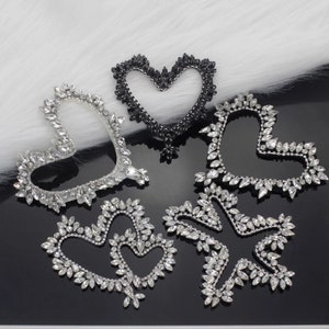 May include: A collection of heart-shaped rhinestone embellishments in silver and black. The sparkling designs vary in size and style, with clear crystals outlining the heart shapes. The embellishments are arranged on a reflective black surface.