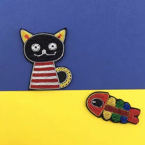 May include: A black cat patch with yellow inner ears, white circular eyes, and a red and white striped body, paired with a multi-coloured fish skeleton patch featuring red, yellow, green, and blue segments, both crafted with detailed beadwork.