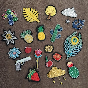 May include: A collection of assorted embroidered patches featuring various designs including a green cactus, a yellow leaf, a golden tree, a silver cloud with a star, a blue anchor, a pineapple, a flower, a handgun, a rose, a cherry, and a peacock feather.