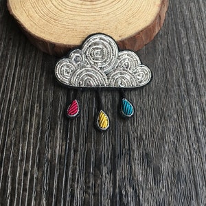 May include: A cloud-shaped brooch featuring intricate silver metallic embroidery and swirling patterns. Three dangling teardrop-shaped charms hang from the bottom, coloured in vibrant red, yellow, and blue, each outlined with black thread.