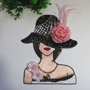 Può includere: Un ricamo a forma di toppa in bianco e nero che raffigura una donna con un cappello nero con un fiore rosa e una piuma. La donna indossa una collana e un fiore rosa sulla spalla.