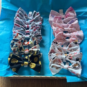 Puede incluir: Una colección de lazos de tela con varios diseños, incluyendo coches, caras sonrientes y motivos florales. Los lazos están dispuestos en filas sobre una superficie azul, con un fondo de madera visible.