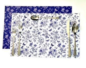 May include: Two rectangular placemats featuring a blue and white floral and butterfly pattern. One placemat has a white background with blue print, while the other has a blue background with white print. Stainless steel cutlery is arranged on top.