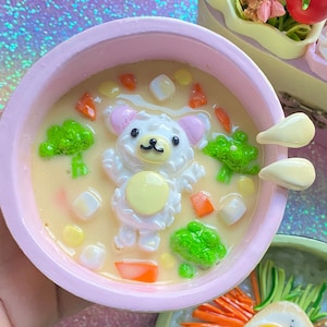 Puede incluir: Primer plano de esculturas de alimentos en miniatura, que incluyen un cuenco rosa de sopa con una figura de oso, brócoli y zanahorias. También se ve un cuenco verde de arroz con verduras y un huevo frito. Un recipiente de arroz y verduras está en el fondo.