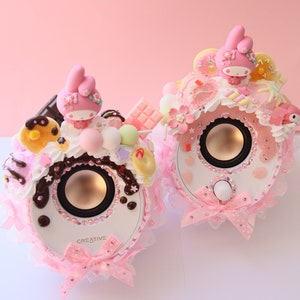 Op de afbeelding: Twee decoratieve luidsprekers met een witte basis, versierd met roze strikken, kant en snoep. Elke luidspreker heeft een cartoonkarakterfiguur en is versierd met strass-steentjes. Het woord "CREATIVE" staat op de luidspreker.