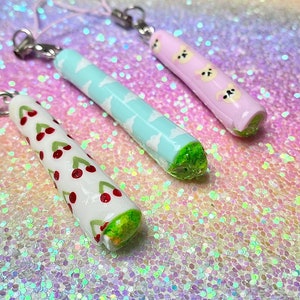 Puede incluir: Tres colgantes decorativos con herrajes plateados. Uno es blanco con cerezas rojas, otro azul claro con nubes blancas y el tercero rosa con caras de oso. Cada colgante tiene una base verde.