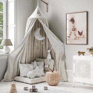 Puede incluir: Una cama con dosel blanco con un m&oacute;vil blanco y dorado de luna y estrellas colgando de la parte superior. Hay almohadas blancas y un conejito de peluche blanco en la cama. Una cesta de mimbre con forma de manzana se encuentra en el suelo. Hay bloques de madera con letras en ellos en el suelo. Una impresi&oacute;n enmarcada de una ni&ntilde;a con un conejito y pollos est&aacute; en la pared.