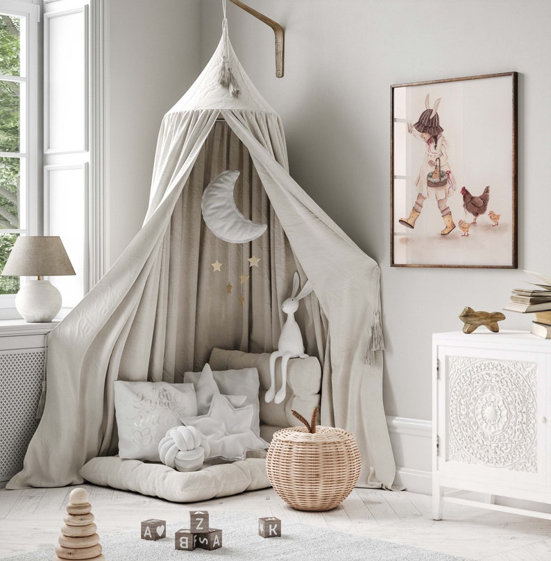 Puede incluir: Una cama con dosel blanco con un m&oacute;vil blanco y dorado de luna y estrellas colgando de la parte superior. Hay almohadas blancas y un conejito de peluche blanco en la cama. Una cesta de mimbre con forma de manzana se encuentra en el suelo. Hay bloques de madera con letras en ellos en el suelo. Una impresi&oacute;n enmarcada de una ni&ntilde;a con un conejito y pollos est&aacute; en la pared.