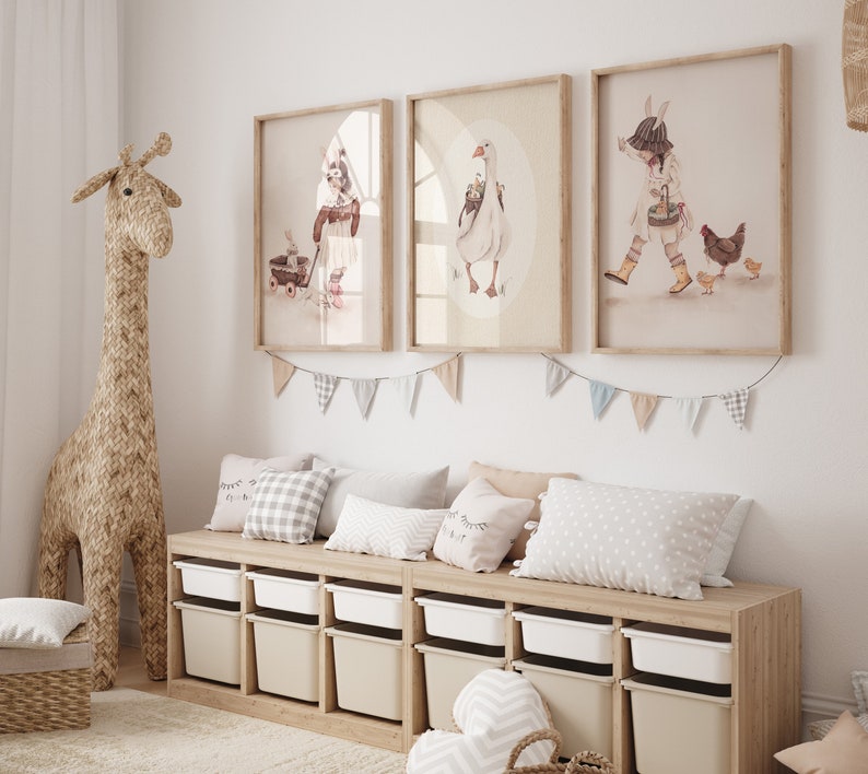 Puede incluir: Una estanter&iacute;a de madera clara con cubos de almacenamiento blancos, un banco de madera clara con tres almohadas y tres ilustraciones enmarcadas de una ni&ntilde;a, un ganso y una ni&ntilde;a con pollos. Las ilustraciones est&aacute;n en un marco marr&oacute;n claro con un paspart&uacute; blanco.