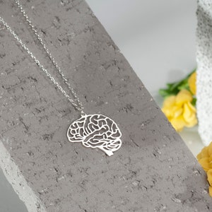 Peut inclure: Un collier en argent avec un pendentif en forme de cerveau. Le pendentif est découpé et présente un design détaillé. Le collier est posé sur une surface texturée grise.