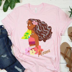 May include: Pink t-shirt with a cartoon image of Moana, a Polynesian princess from the Disney movie Moana. The image shows Moana with long brown hair, a flower in her hair, and a necklace. The text "Moana" is printed below the image.