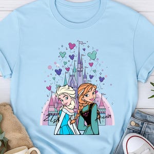 May include: Light blue t-shirt featuring a graphic of two animated princesses in front of a castle, with heart and Mickey Mouse-shaped balloons. The princesses are Elsa and Anna. The shirt is paired with jeans and white sneakers.